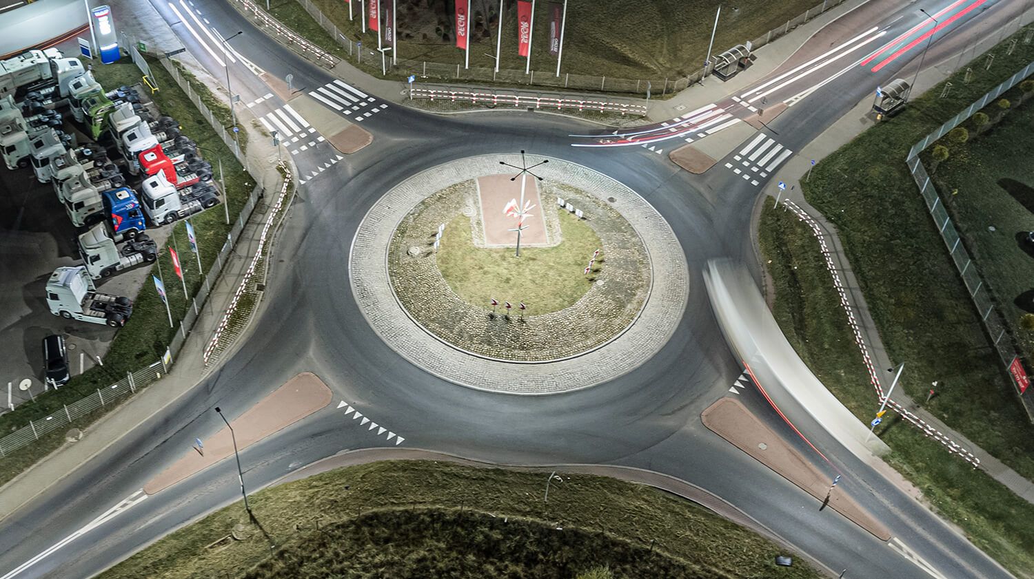 Vue aérienne nocturne d'un rond-point éclairé par des lampadaires, montrant la circulation automobile et la signalisation indiquant le sens de circulation.